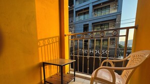 A balcony with a small table and a chair, a metal railing, and a view of a building with balconies. - Stay At Bedroom (Patong)