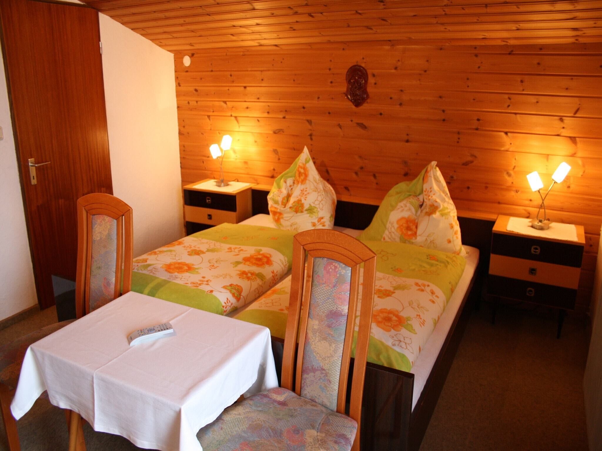 Una habitación con paneles de madera, una cama, dos mesitas de noche, una mesa con una silla y una puerta.