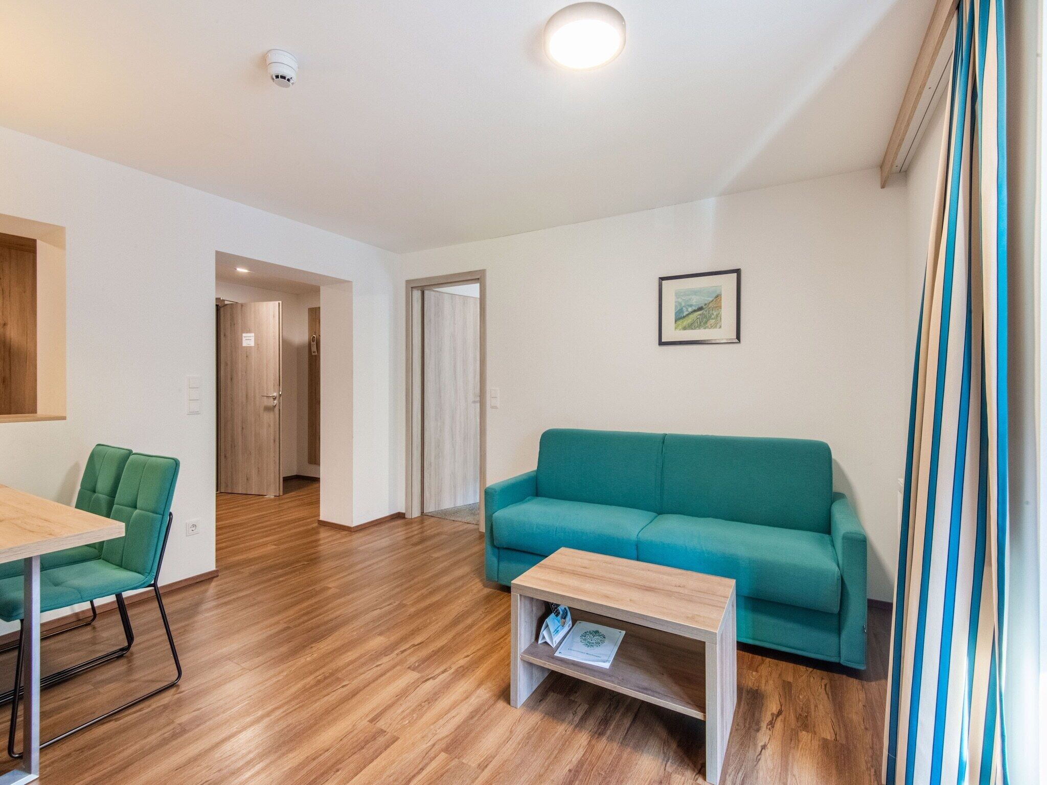 A modern living room with a teal sofa, a wooden coffee table, and a dining area with a green chair.