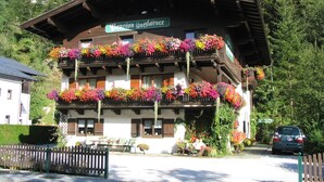 Ein traditionelles Holzhaus mit Balkonen, die mit bunten Blumen geschmückt sind, einem Schild, das es als Pension kennzeichnet, und einem geparkten Auto in der Nähe.