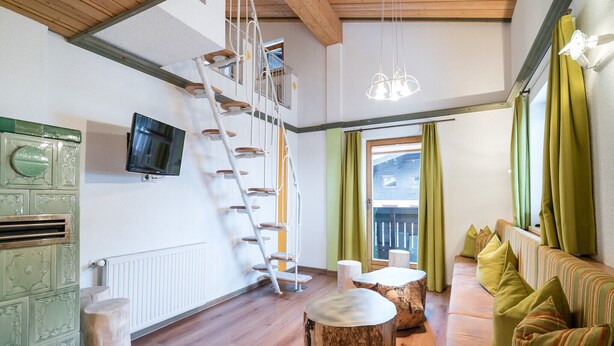 Ein Zimmer mit Holzboden, einem Schlafplatz in der Schlafloge, einem Fernseher, einer Holzleiter und einem grünen Sofa.