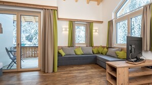 A cozy living room with a wooden floor, a gray sectional sofa with green cushions, a wooden TV stand, and large windows with curtains. - Chalet in Saalbach near Ski Lift (Saalbach-Hinterglemm)
