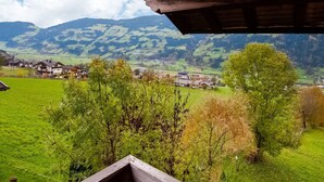 A view from a wooden balcony overlooking a green valley with houses and mountains in the background.