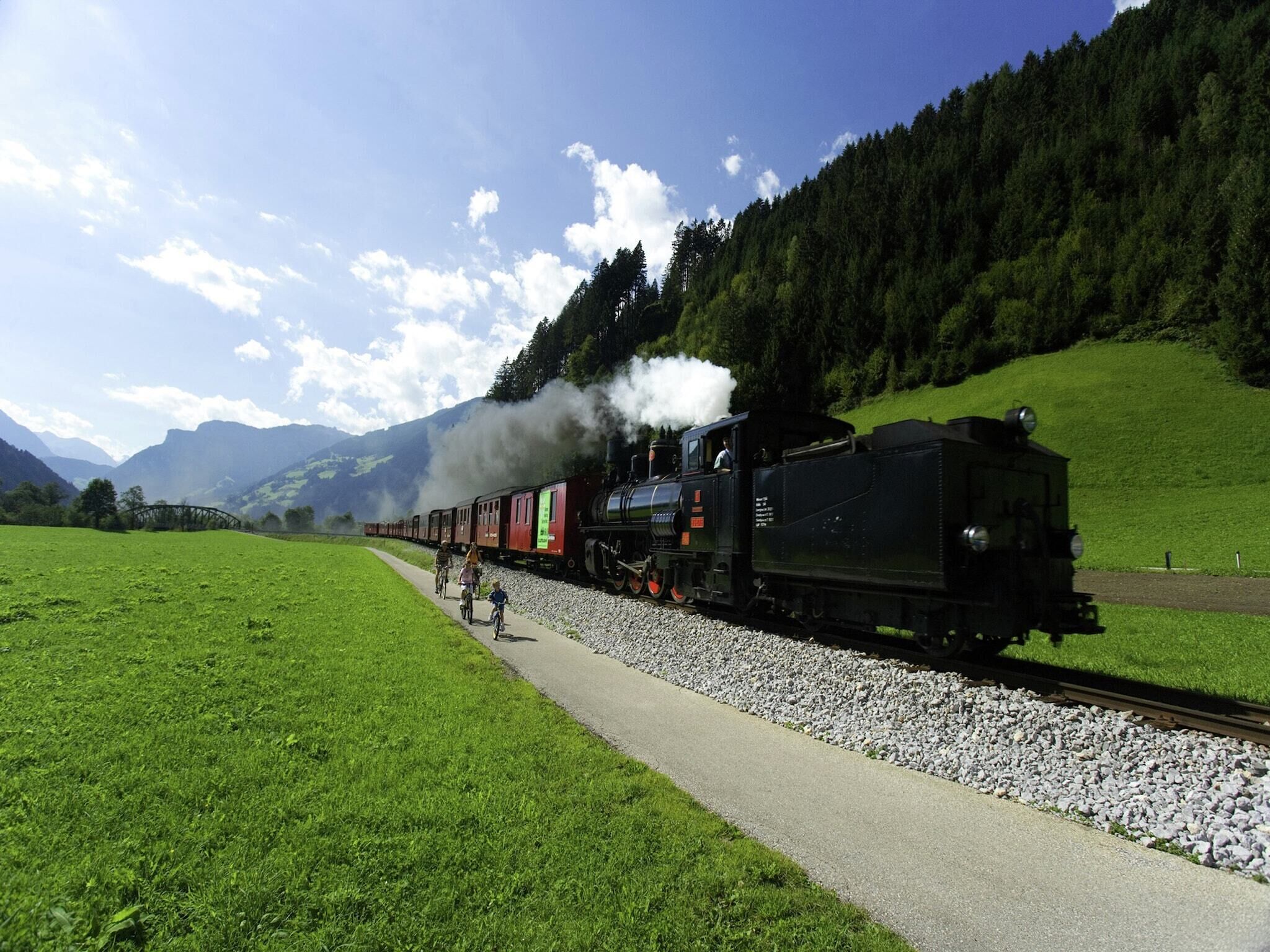A steam locomotive with passengers on a railway track surrounded by green fields and mountains.
