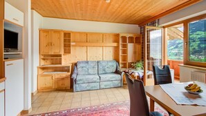 A cozy room with a wooden ceiling, a sofa, a dining table with a bowl of fruit, and a view of green hills through large windows.
