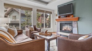 A cozy living room with leather sofas, a fireplace, and a large window overlooking a backyard. - Spacious Resort Condo | Near Banff (Canmore)