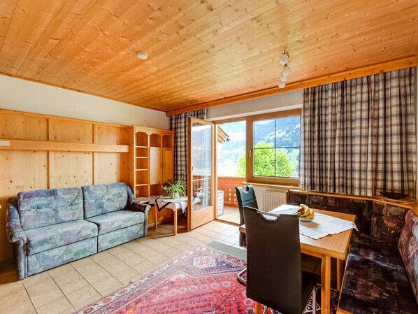 A cozy room with a wooden ceiling, a sofa, a dining table with chairs, and a view of greenery outside.