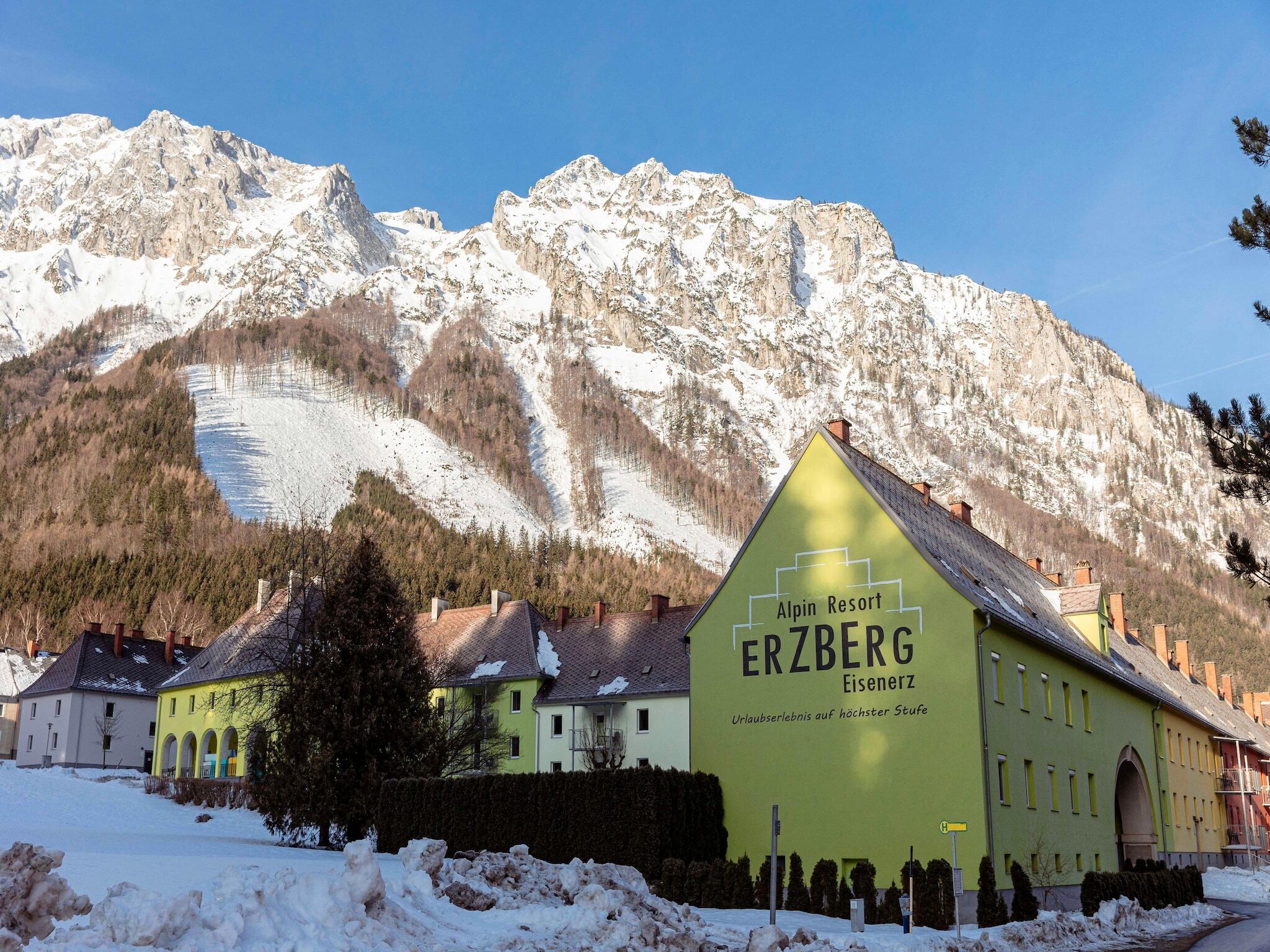 A mountain resort building with a sign for "Alpin Resort ERZBERG Eisenerz" and a view of snow-covered mountains in the background.