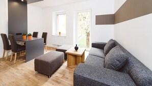 A modern living room with a grey sofa, a wooden coffee table, and a dining area with a table and chairs. There are two windows providing natural light. - Apartment in Styria with bubble bath (No Deposit) (Eisenerz)