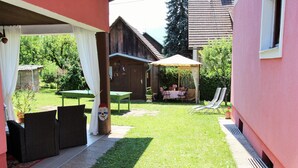 Ein überdachter Außenbereich mit Tisch und Stühlen, einer Rasenfläche und einem Holzgebäude im Hintergrund.
