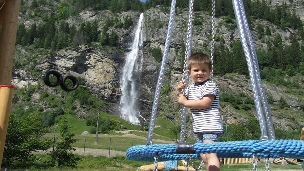 Niño en un columpio con una cascada y montañas rocosas al fondo.
