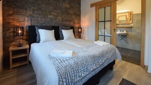 Une chambre avec un mur en pierre, un lit avec une literie à rayures, une table de chevet, un plancher en bois et une salle de bain avec un lavabo et un miroir.