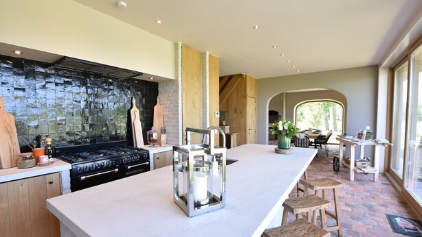 A modern kitchen with a large island, a built-in stove, and a stone backsplash. There is a dining area with a table and chairs visible through an archway. - Beautiful Holiday Home with Terrace, Garden (Diksmuide)