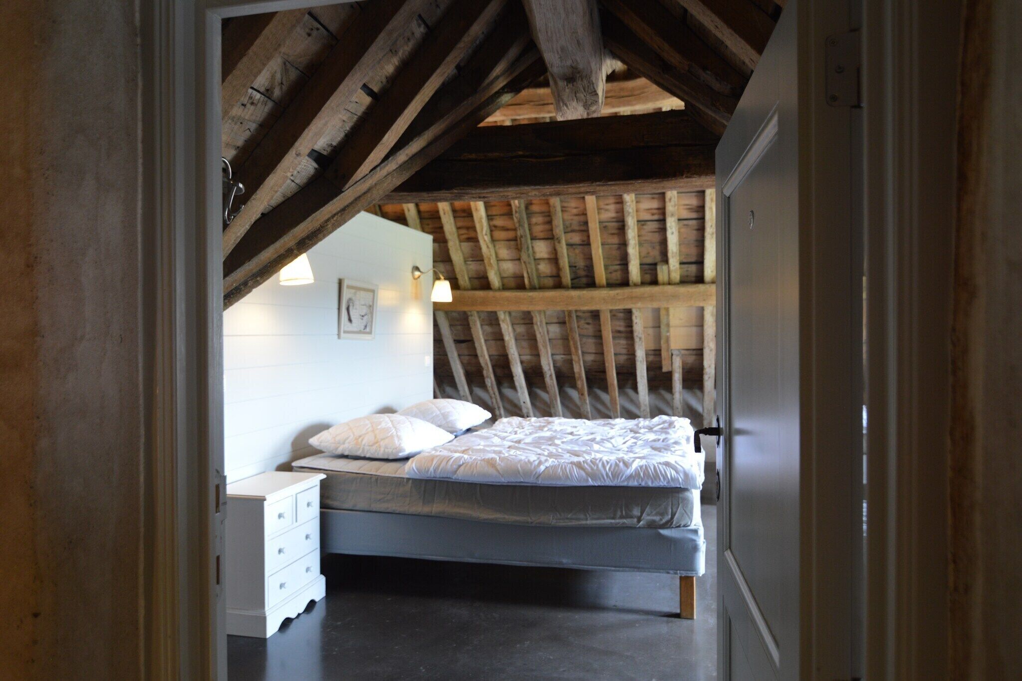 A bedroom with a wooden ceiling, a bed with white bedding, a nightstand, and a wall-mounted light fixture.