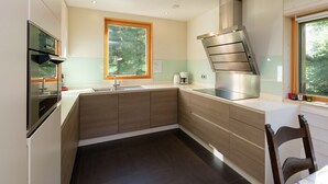 A modern kitchen with a built-in oven, a stainless steel range hood, a window with a view of trees, and a dark tiled floor.