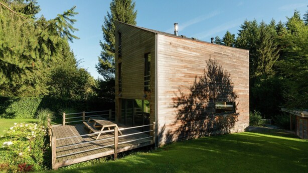 A modern wooden cabin with a deck and a chimney, surrounded by lush greenery.