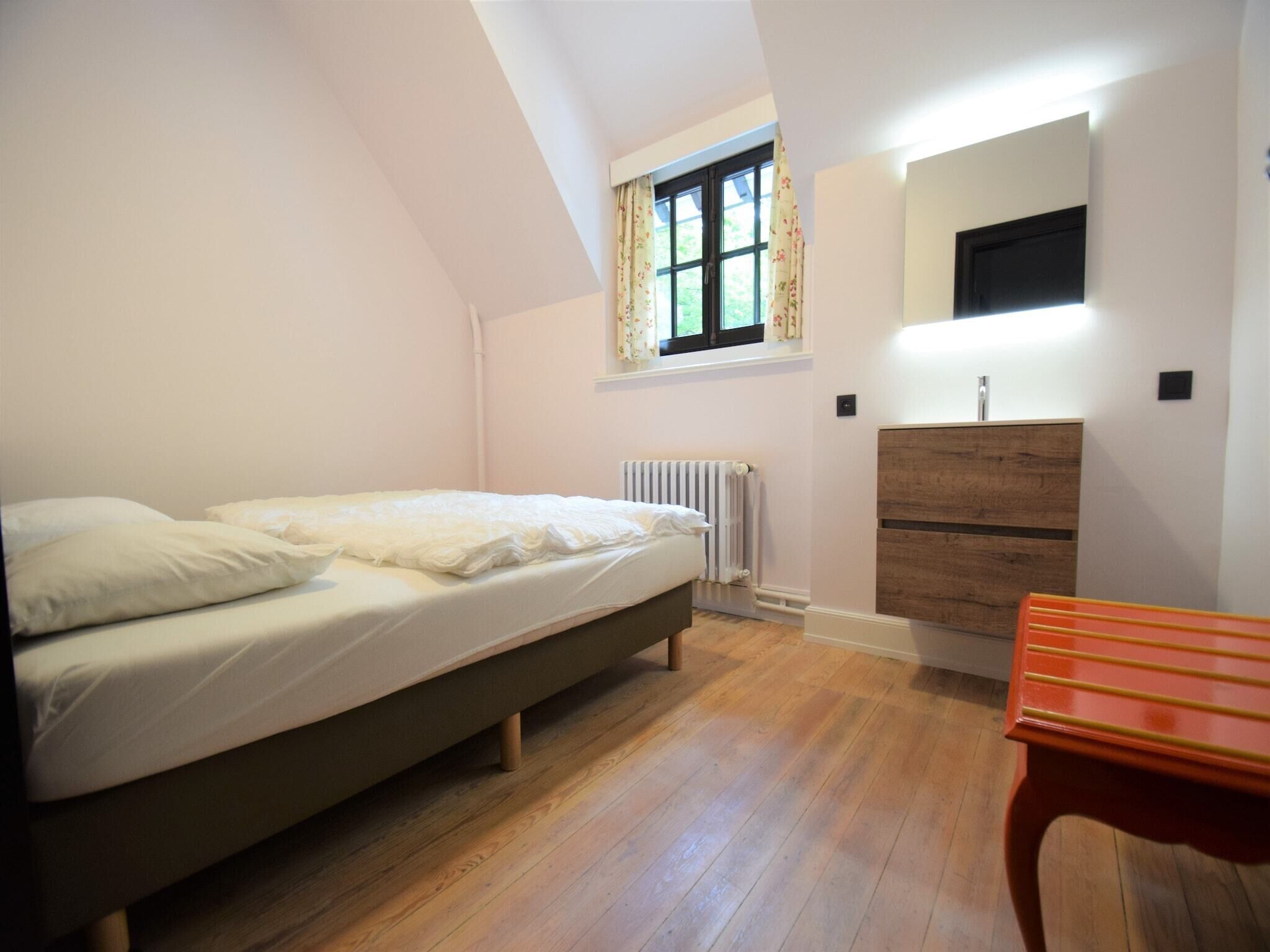 Une chambre à coucher avec un lit, une fenêtre avec des rideaux, un radiateur, une commode en bois et une petite table.