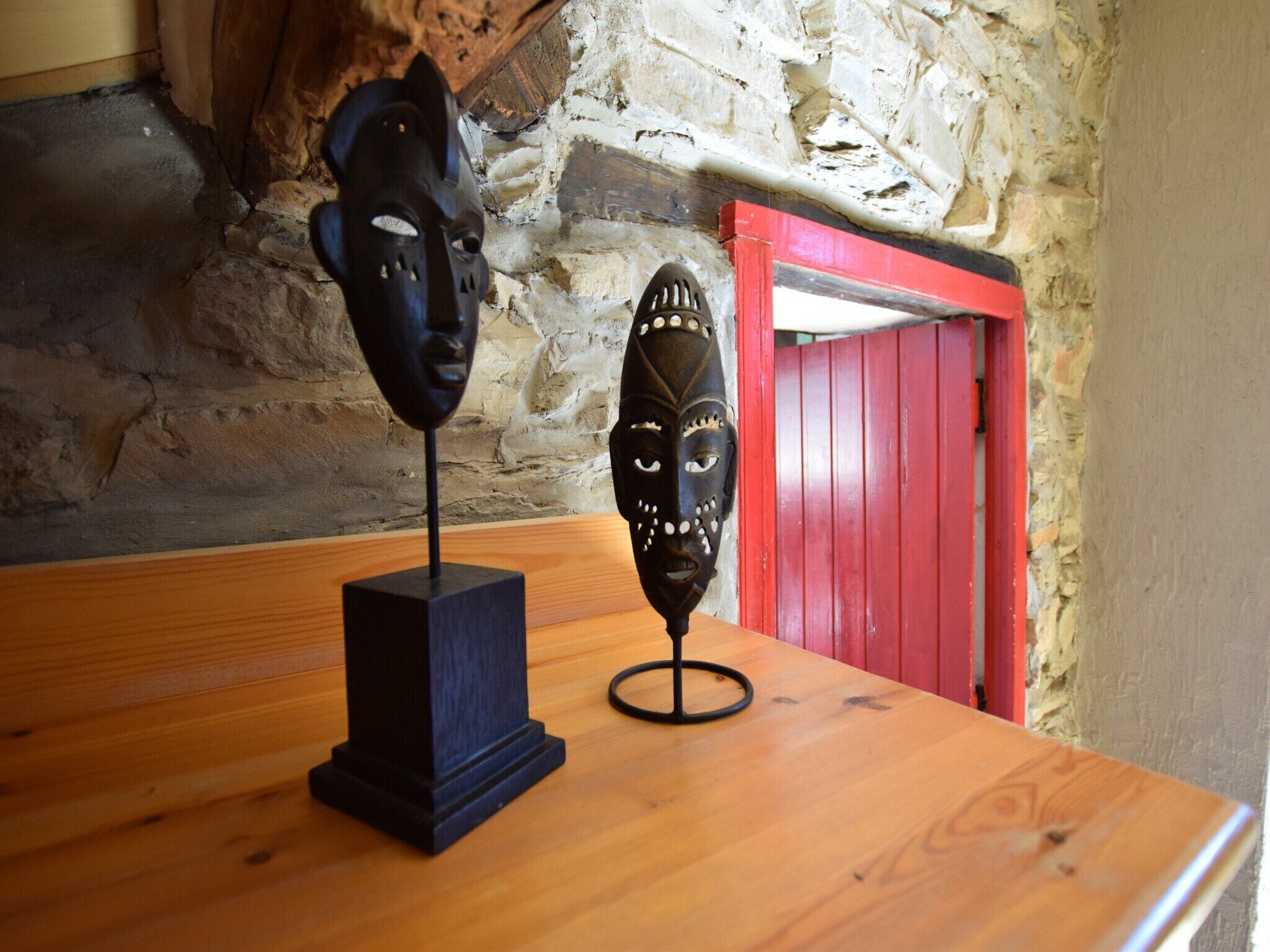 Dos mesas de madera con esculturas negras en una habitación con paredes de piedra y una puerta roja.