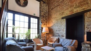 A cozy lounge area with wicker furniture, a fireplace, and large windows allowing natural light. - Gouvy Forest Farmhouse (Gouvy)
