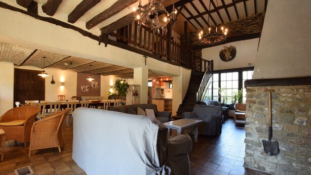 A spacious interior with a high ceiling, exposed wooden beams, a stone fireplace, and a cozy seating area with wicker furniture. - Gouvy Forest Farmhouse (Gouvy)