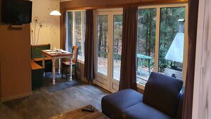A cozy living room with a sofa, a dining table with chairs, and a television. Large windows offer a view of the forest.