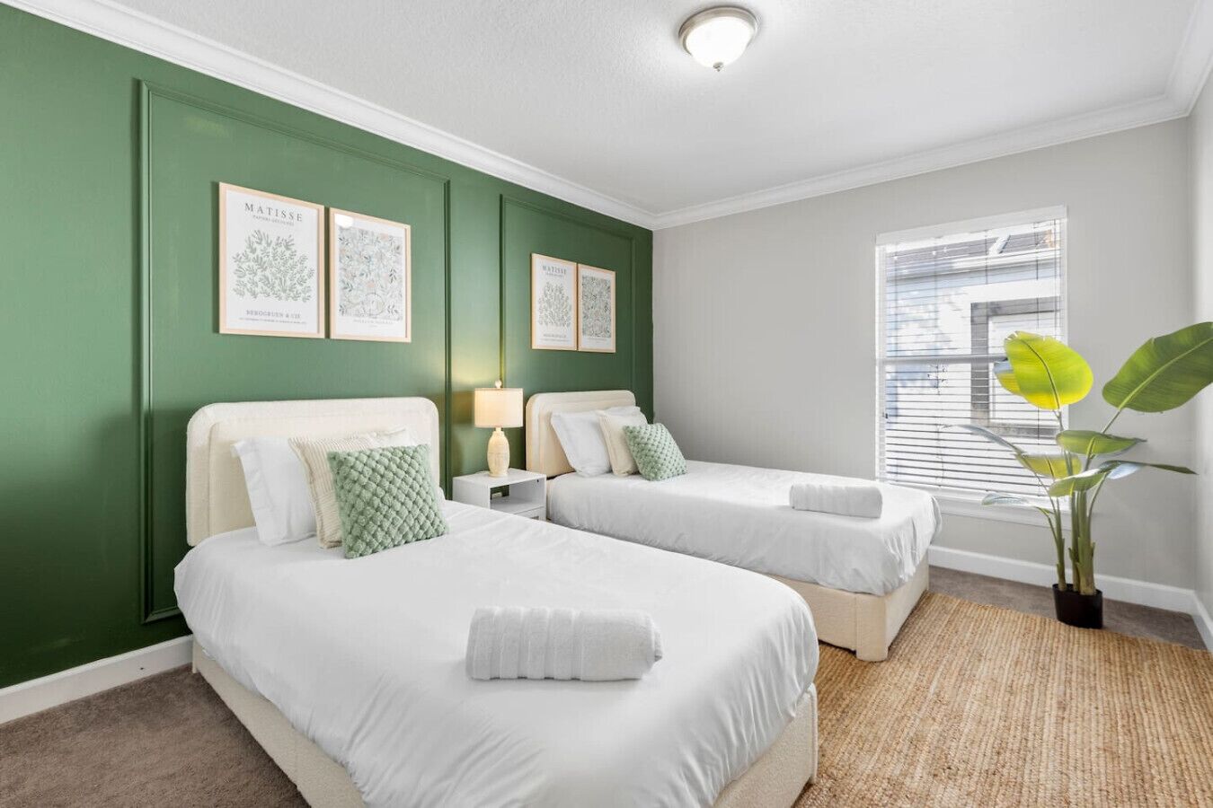 A bedroom with two beds, green walls, framed artwork, a window, and a plant.