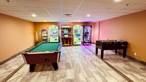 Mesa de bilhar, mesa de futebol de mesa e máquinas de fliperama em um espaço com piso ladrilhado e teto alto.