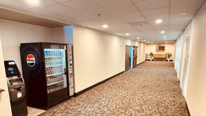 Corredor de hotel com piso coberto por carpete, uma máquina de refrigerantes Pepsi e um balcão de recepção.