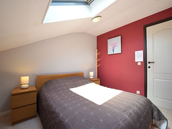 A bedroom with a skylight, a bed with a patterned bedspread, a nightstand with a lamp, a wooden headboard, and a red accent wall.