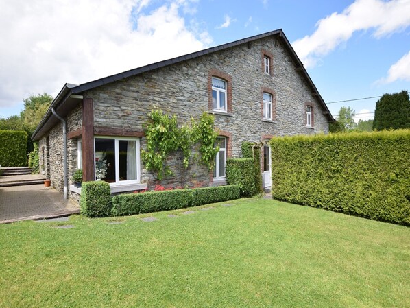 A stone house with a sloped roof, a front door, and a small garden area with a hedge and a tree.