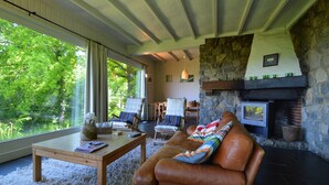 A cozy living room with a stone fireplace, leather sofa, and a large window overlooking greenery. - Modern chalet with a view (Humain)