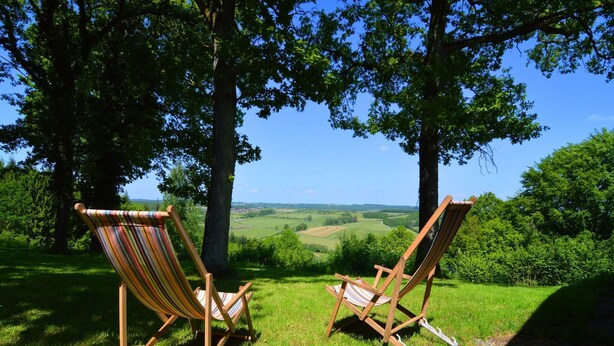 Two wooden deck chairs with striped cushions, positioned on a grassy slope overlooking a scenic landscape with trees and a clear blue sky. - Modern chalet with a view (Humain)