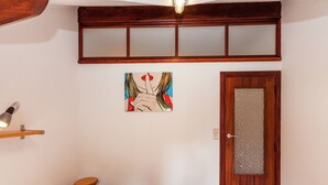 Un dormitorio con cama, una puerta de madera con panel de vidrio, un taburete pequeño, un estante en la pared y una lámpara en el techo.