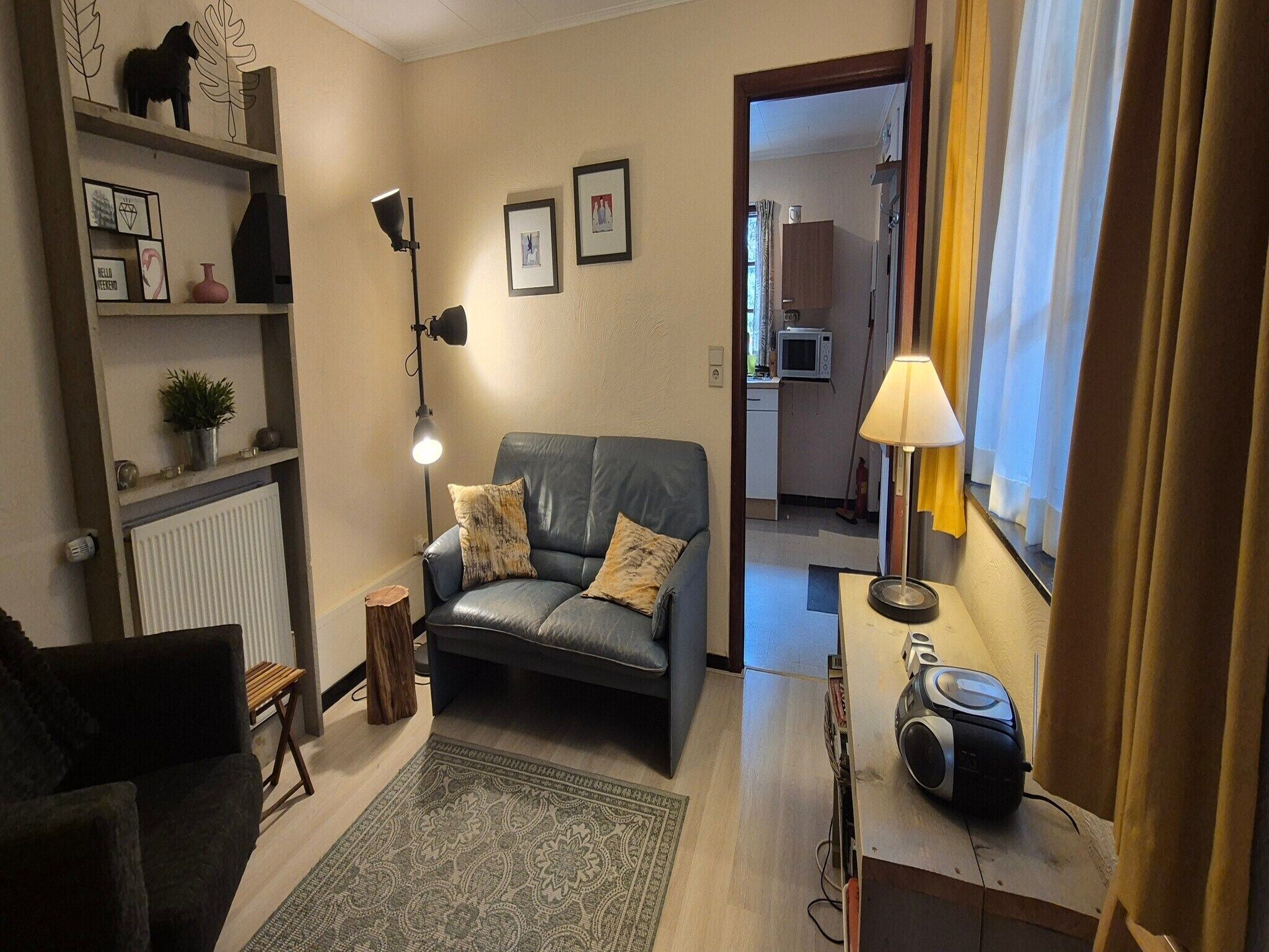 A living room with a sofa, a lamp, a small table with a radio, and a view into the kitchen.