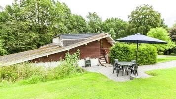 A wooden cabin with a sloped roof, a small porch, and a table with chairs under a blue umbrella.