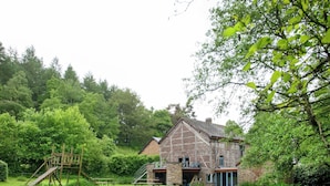 Un bâtiment de style rustique, évoquant une grange, avec des fondations en pierre et une structure supérieure en bois, entouré d’une végétation luxuriante et agrémenté d’une structure de jeu en bois.