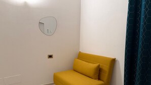Un piccolo divano giallo in un angolo della stanza, con una parete bianca, uno specchio rotondo e una tenda blu scuro.