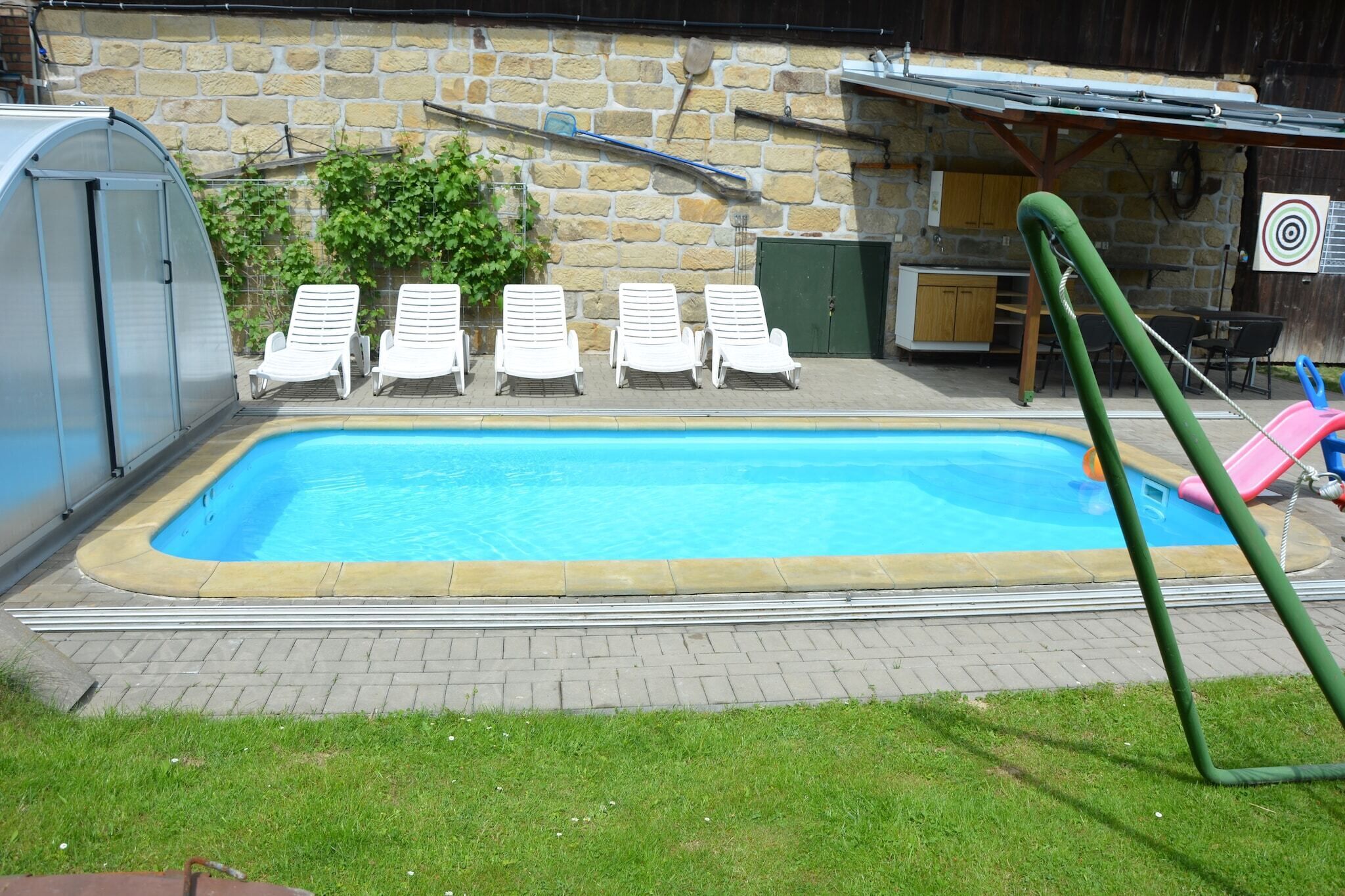 Una piscina con sillas de descanso, un invernadero y un muro de piedra.