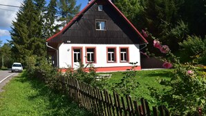 A two-story house with a red roof and white walls, surrounded by greenery and a wooden fence.