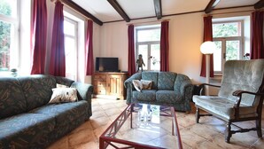 A living room with a sofa, armchair, and a glass coffee table. There is a television on a wooden cabinet and a window with curtains.