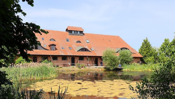 A large brick building with a red-tiled roof, surrounded by a pond and greenery. - Ferienwohnung Im Heumond in Buschhagen (Buschenhagen)