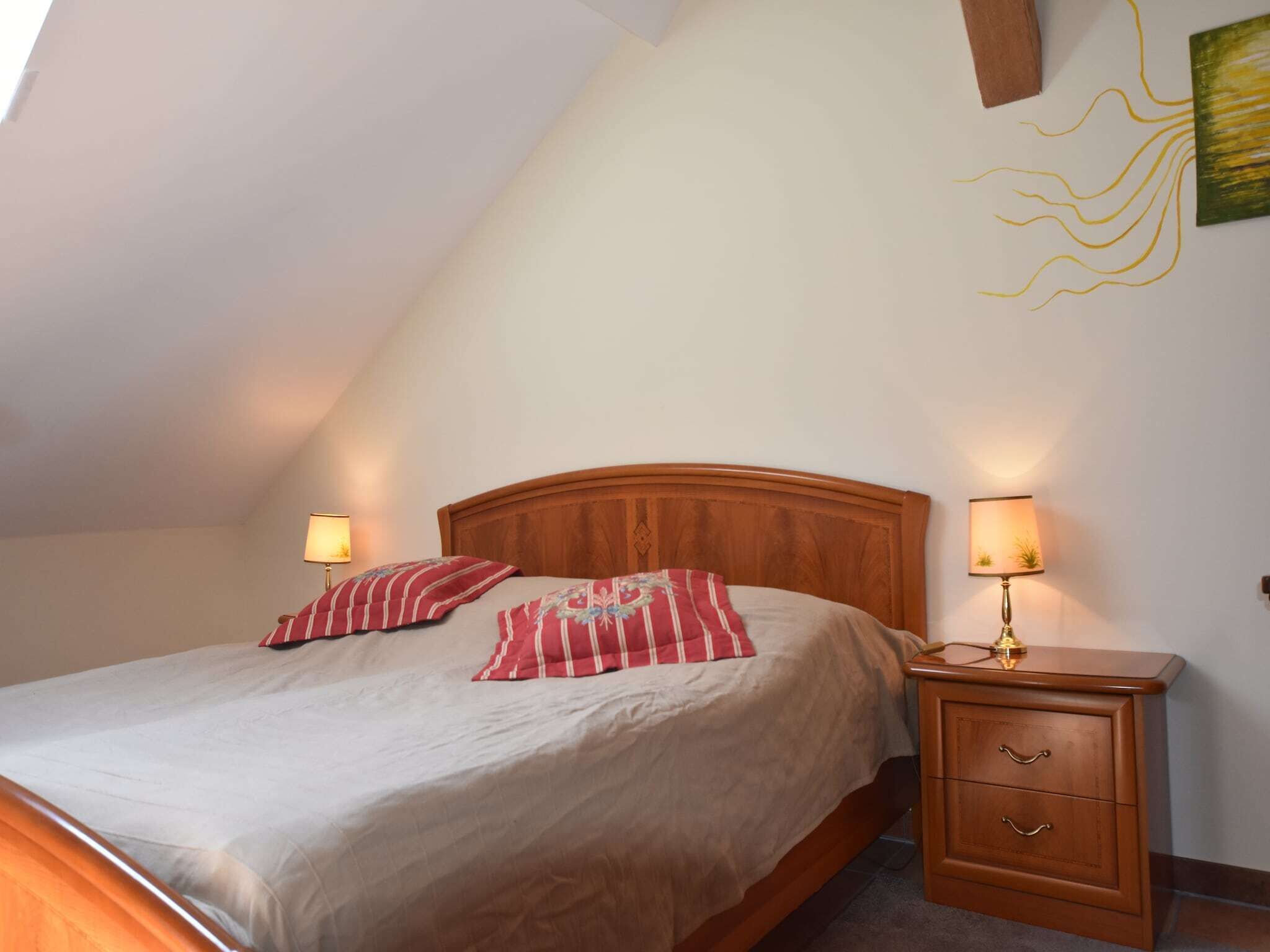 Un dormitorio con techo inclinado, una cama de madera con dos almohadas, una mesita de noche con una lámpara y un cuadro en la pared.