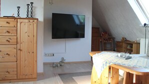 A room with a wooden cabinet, a flat-screen TV mounted on the wall, a small table with a tablecloth, and a window with a view of the outside. - Moderne Ferienwohnung in Meeresnähe in Tarnewitz (Boltenhagen (Ostseebad))
