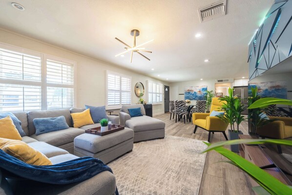 A spacious living room with a grey sectional sofa, a yellow armchair, and a blue bean bag. There is a dining area with a zebra-striped table and chairs, and a kitchen in the background.