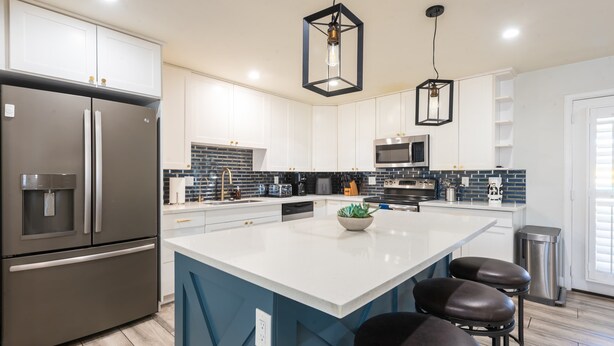 A modern kitchen with white cabinets, a stainless steel refrigerator, a large island with stools, and hanging pendant lights. - Home with Backyard Fun: Hot Tub, Golf, BBQ, Games! (Phoenix)