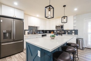 A modern kitchen with white cabinets, a stainless steel refrigerator, a large island with stools, and hanging pendant lights.