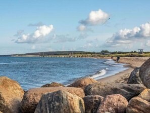 A beach with large rocks in the foreground, a pier extending into the water, and a few people walking along the shore.