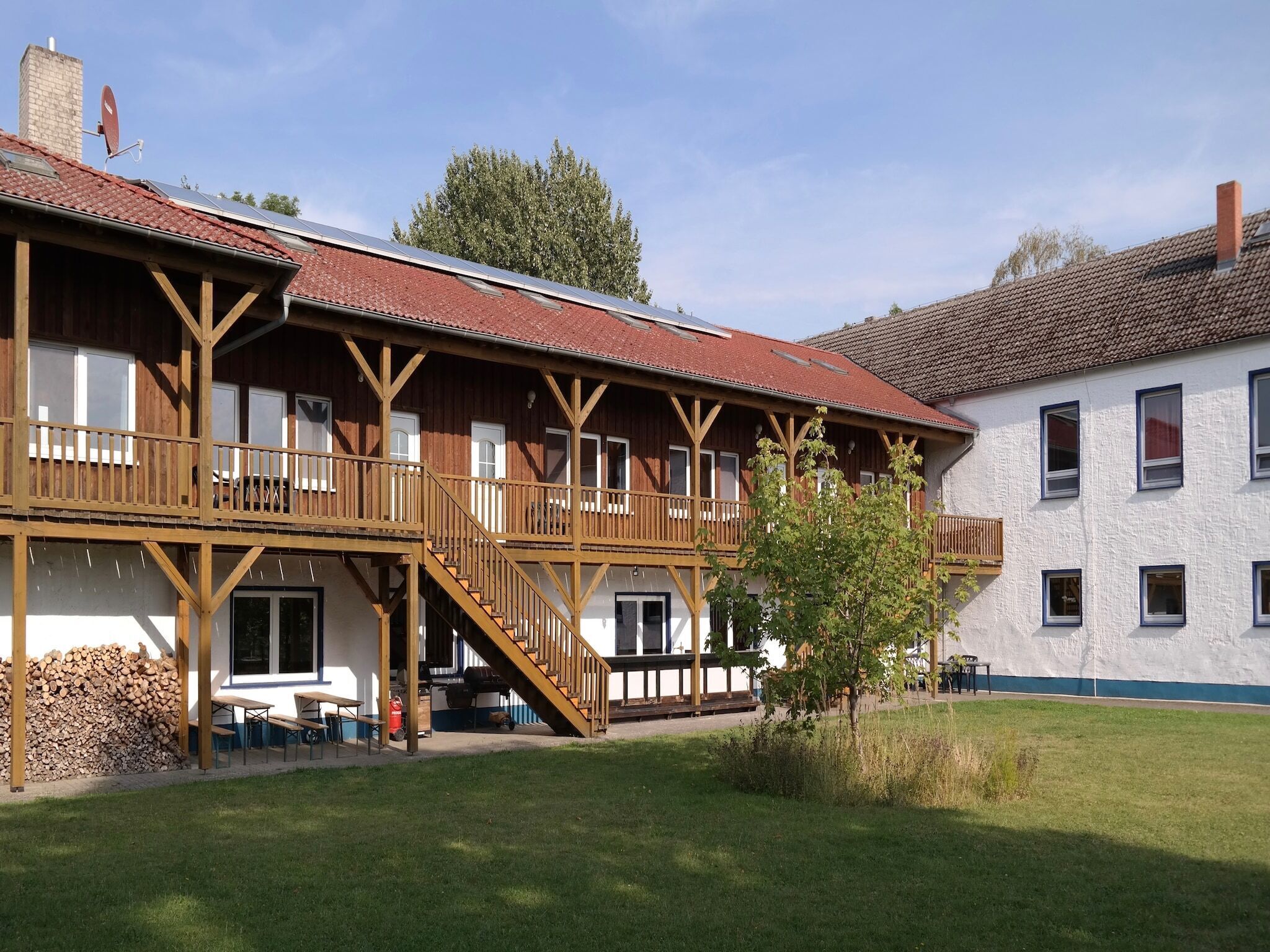 Un bâtiment en bois à deux étages, doté de balcons, d’un espace extérieur couvert aménagé en coin salon et d’une cour verdoyante.