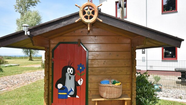Une cabane en bois avec une porte rouge, un gouvernail de bateau et un panier posé sur un banc.