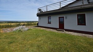 A single-story building with a red door and windows, a small balcony with a railing, and a grassy area in front. - Ferienwohnung in Bastorf for 3 Personen (Bastorf)